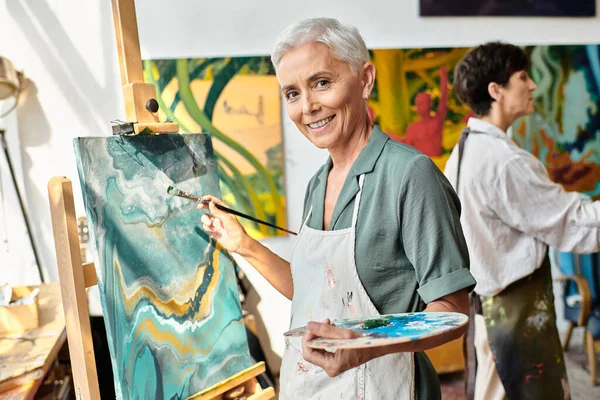 Sonriendo mujer madura pintura en caballete y sonriendo a la cámara en taller de arte, proceso creativo - foto de stock