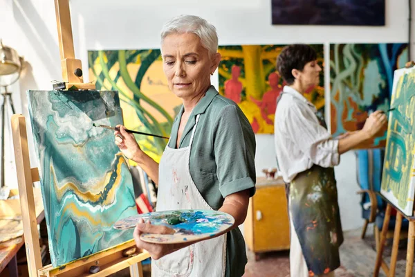 Talented middle aged woman with palette painting on easel near female friend in art workshop — Stock Photo