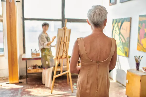Элегантная зрелая модель, позирующая рядом с художницей, рисующая на мольберте в художественной мастерской, творческий процесс — стоковое фото
