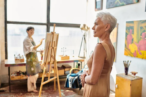 Стильная зрелая модель, позирующая рядом с художницей, рисующая на мольберте в художественной мастерской, творческий процесс — стоковое фото