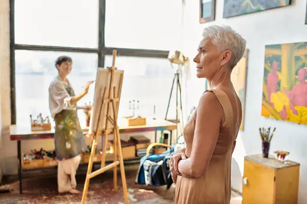 Elegante modelo maduro posando perto artista feminino dor no cavalete na oficina de arte, processo criativo — Fotografia de Stock