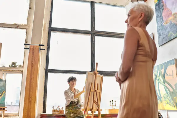 Modèle élégant d'âge moyen posant près de peinture d'artiste féminine en atelier, processus artistique — Photo de stock