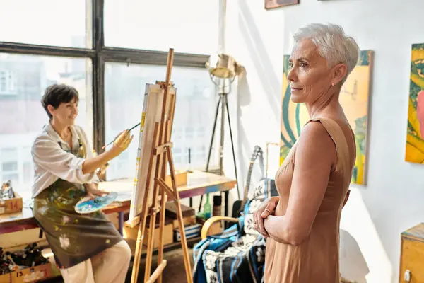 Elegant middle aged model posing near female artist painting in workshop, creative process — Stock Photo