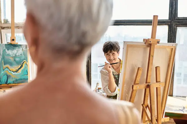 Gioiosa artista femminile parlando con offuscata modello maturo in posa in laboratorio artigianale, processo creativo — Foto stock