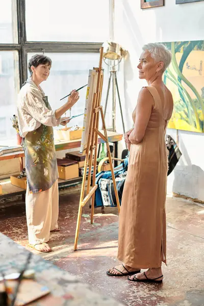 Talented female artist painting mature model in elegant dress in art workshop, artistic process — Stock Photo