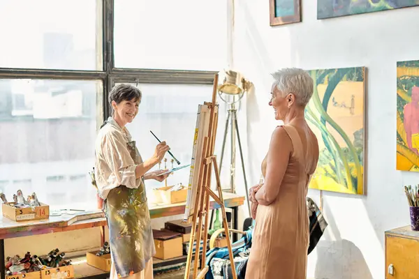 Счастливая зрелая женщина рисует элегантную подругу, позирующую в художественной мастерской, художественный процесс — стоковое фото