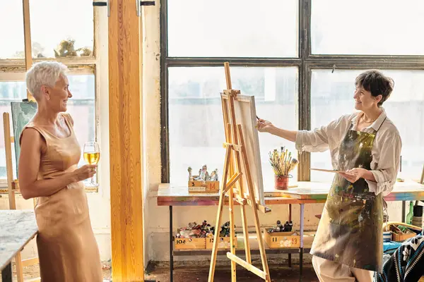 Счастливая зрелая художница рисует элегантную подругу, позирующую с бокалом вина в художественной студии, творчество — стоковое фото