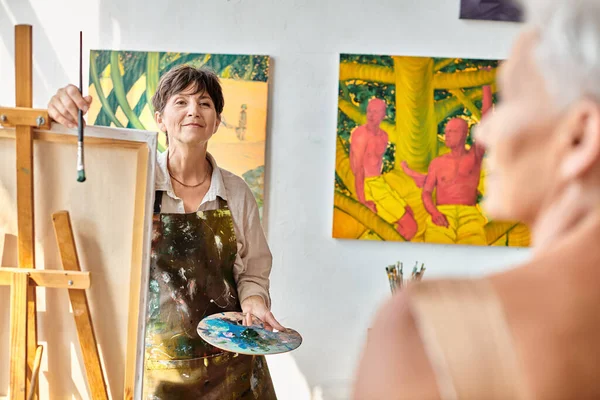Heureux artiste femme regardant flou modèle mature posant dans l'atelier d'art, processus créatif — Photo de stock