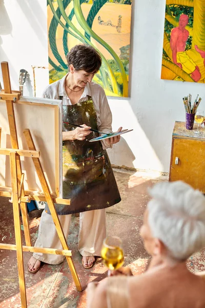 Cheerful mature artist painting blurred model with wine glass in art workshop, creative process — Stock Photo