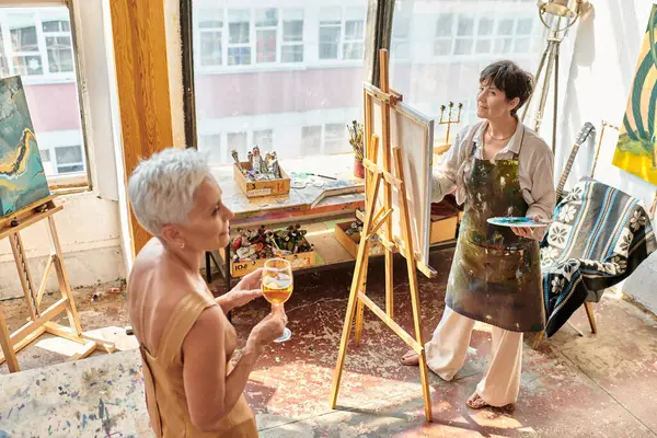 Inspired mature artist painting graceful model with wine glass in art workshop, creative process — Stock Photo