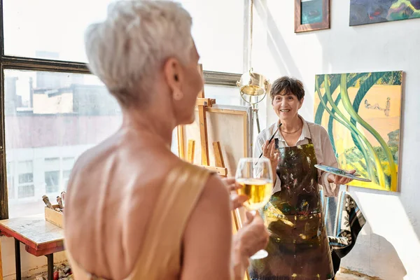 Felice artista maturo pittura modello femminile offuscata con bicchiere di vino in laboratorio d'arte, processo creativo — Foto stock