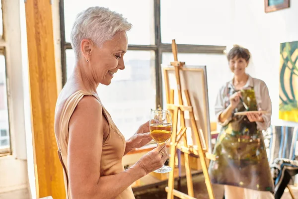 Smiling middle aged model posing with wine glass near blurred female artist in art workshop — Stock Photo