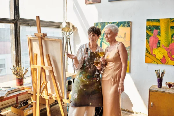 Mature female artist with elegant model holding wine glasses and looking at easel in art studio — Stock Photo