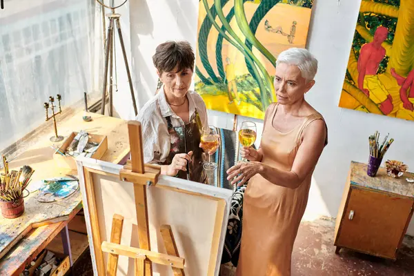 Middle aged model with female artist holding wine glasses and looking at easel in art studio — Stock Photo