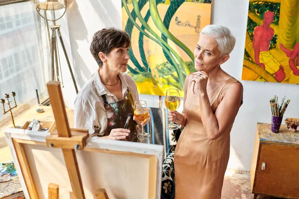 Thoughtful mature model with wine glass looking at easel near professional female artist in studio — Stock Photo