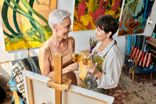 Alegres amigas maduras con copas de vino cerca del caballete en el estudio de arte, artista y modelo de trabajo en equipo - foto de stock