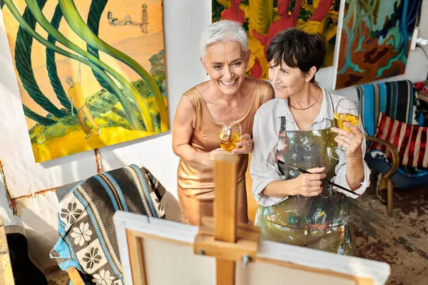 Heureux mature copines avec des verres à vin souriant près de chevalet dans l'atelier, artiste et modèle — Photo de stock