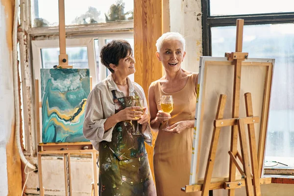 Impressed mature model with wine glass looking at easel near female artist in workshop, artwork — Stock Photo