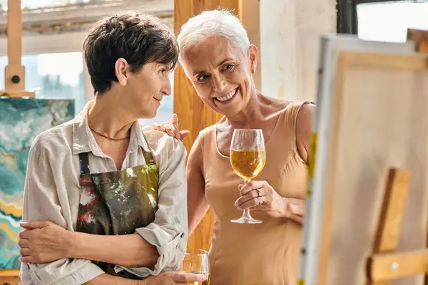 Весёлая зрелая модель с бокалом вина, смотрящая на мольберт рядом с художницей в мастерской — стоковое фото