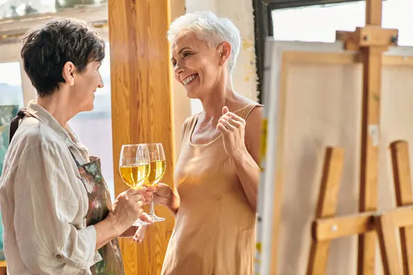 Pleased female painter and elegant mature model clinking wine glasses in modern art workshop — Stock Photo