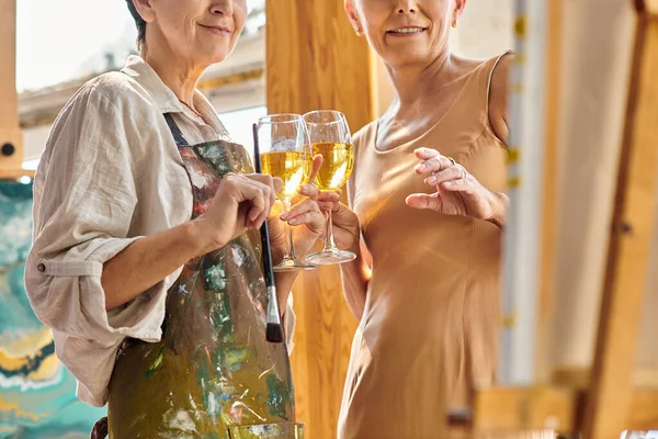 Cropped view of female painter and mature model with wine glasses near easel in modern art workshop — Stock Photo
