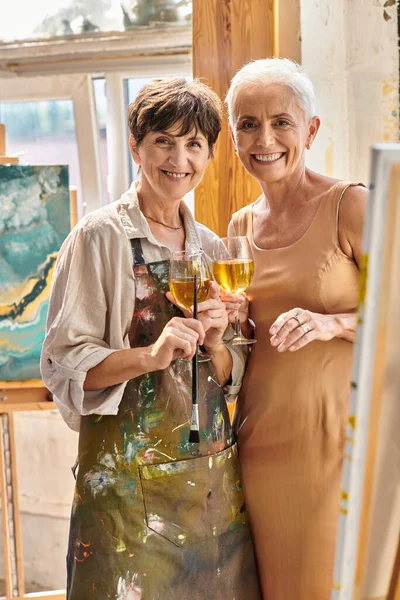 Delighted female painter and stylish mature model with wine glasses looking at camera in workshop — Stock Photo