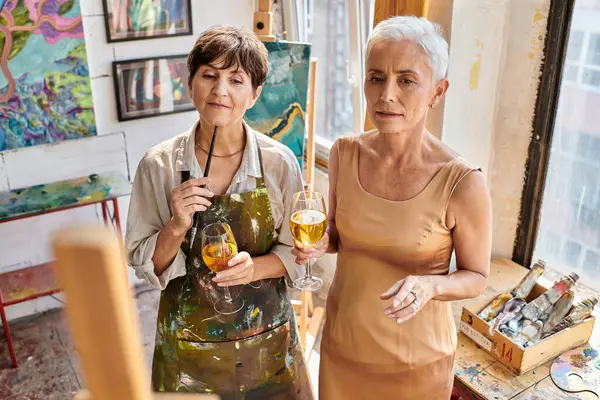 Thoughtful woman artist and stylish mature model standing with wine glasses  in craft workshop — Stock Photo