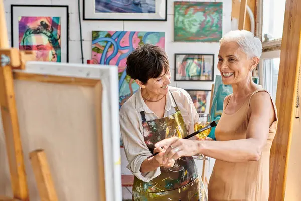 Cheerful and stylish mature model with wine glass pointing at easel near woman painter in art studio — Stock Photo
