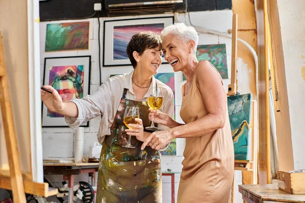 Excited mature woman artist and elegant model with wine glasses laughing near easel in art studio — Stock Photo