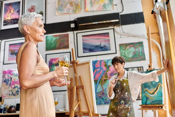 Modelo maduro elegante con copas de vino posando cerca de la mujer pintor en el estudio de arte, proceso creativo - foto de stock