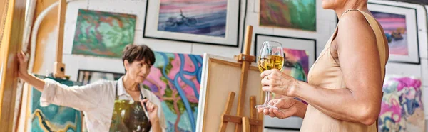 Middle aged model with wine glasses posing near woman painter in art studio, horizontal banner — Stock Photo
