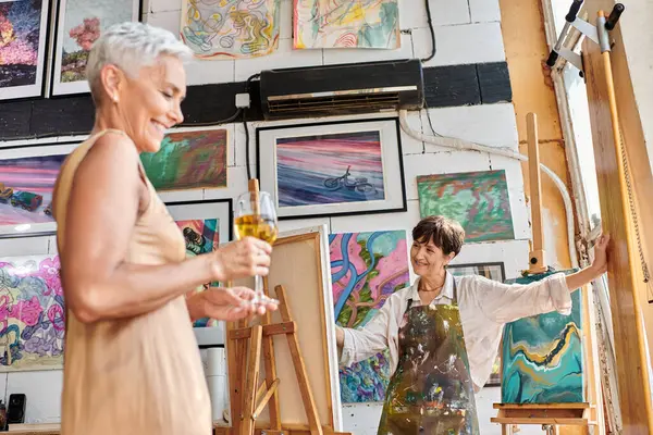 Sourire modèle mature avec des verres à vin posant près de la femme artiste en atelier d'art, processus créatif — Photo de stock