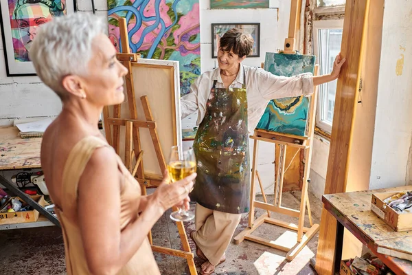 Artista mulher feliz olhando elegante modelo de meia-idade posando com copo de vinho em oficina de arte — Fotografia de Stock