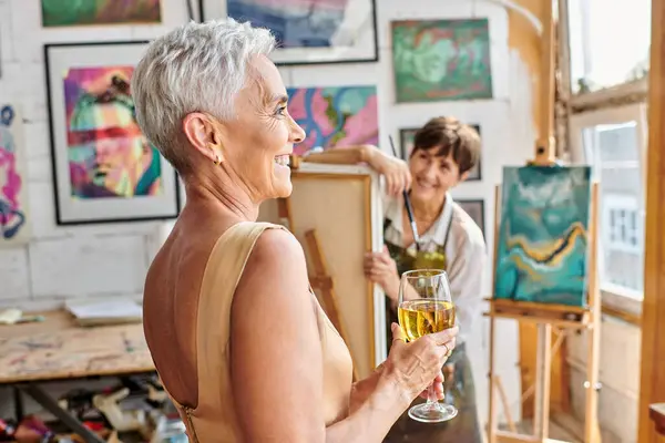 Радісна і стильна жінка позує з келихом вина біля жінки-художника в майстерні, творчий процес — стокове фото