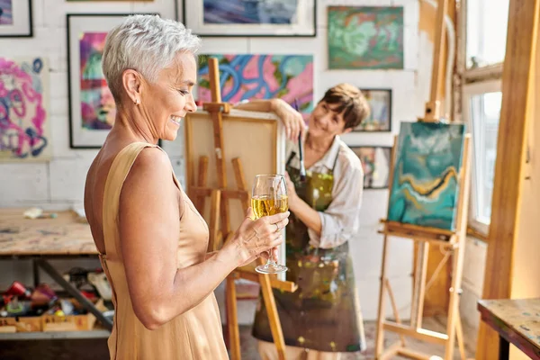 Happy and elegant woman posing with wine glass near female artist in workshop, creative process — Stock Photo