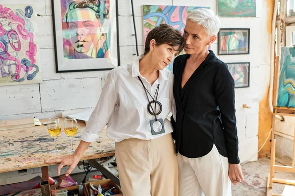 Élégant couple lesbienne mature debout près de peintures colorées dans l'atelier d'art moderne, ensemble — Photo de stock