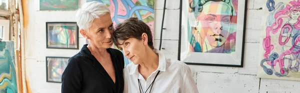 Couple lesbien mature à la mode debout près de peintures dans l'atelier d'art moderne, bannière horizontale — Photo de stock