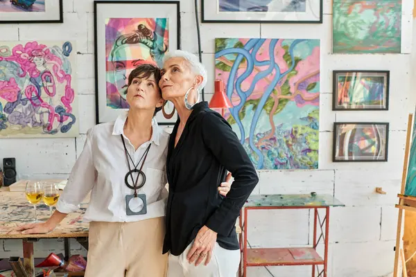 Couple lesbien mature à la mode regardant des peintures colorées dans l'atelier d'art moderne, ensemble — Photo de stock