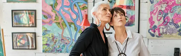 Stylish middle aged lesbian couple looking at creative paintings in modern art workshop, banner — Stock Photo