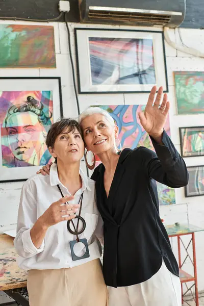Smiling woman pointing with hand near lesbian partner and colorful paintings in art workshop — Stock Photo