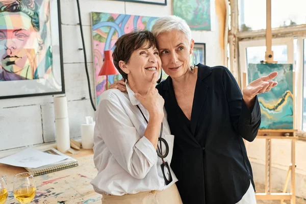 Stilvolle Künstlerin zeigt mit der Hand auf fröhliche lesbische Frau und Gemälde in Kunstwerkstatt — Stockfoto