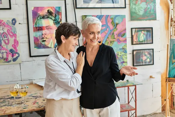 Trendy female artist pointing with hand near joyful lesbian partner and paintings in art workshop — Stock Photo
