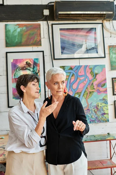 Couple lesbien créatif de même sexe parlant près de peintures colorées dans l'atelier d'art moderne, travail d'équipe — Photo de stock
