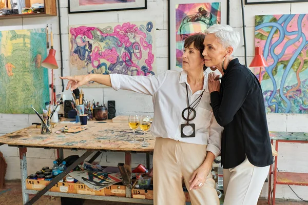 Mature female artist pointing with hand near painting and lesbian partner in modern craft workshop — Stock Photo