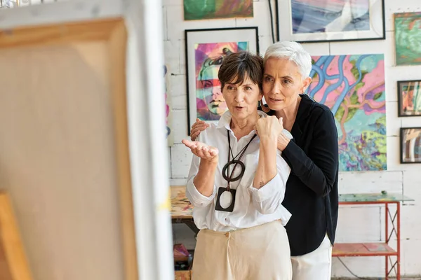Впечатленная художница средних лет, указывающая рукой на партнера-лесбиянку в студии современного искусства — стоковое фото