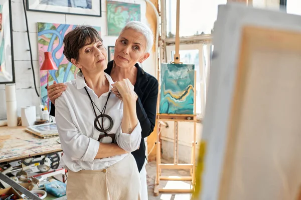 Artistes lesbiennes élégantes et réfléchies regardant la peinture dans l'atelier d'artisanat moderne, l'amour et l'art — Photo de stock