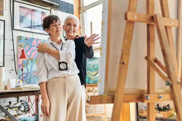 Joyeuse artiste féminine pointant vers chevalet près partenaire lesbienne dans l'atelier, le travail d'équipe et l'art — Photo de stock
