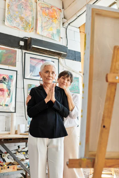 Impressed female artist looking at easel near lesbian partner in workshop, teamwork and art — Stock Photo