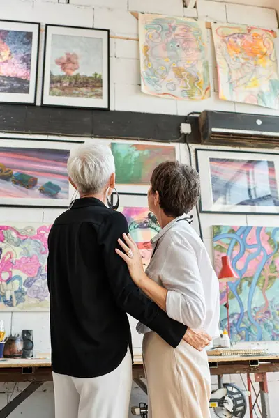 Back view of lesbian couple of artists looking at creative vibrant paintings in modern art workshop — Stock Photo