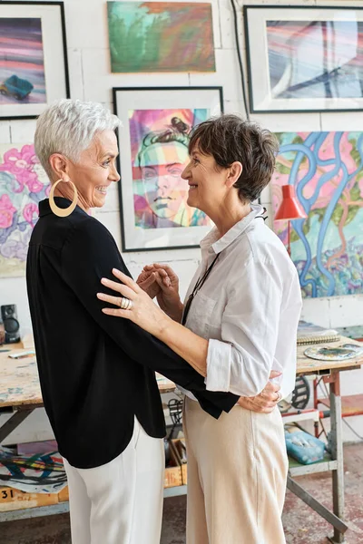 Vista lateral de alegre e elegante casal lésbico de artistas olhando uns para os outros na oficina de arte — Fotografia de Stock